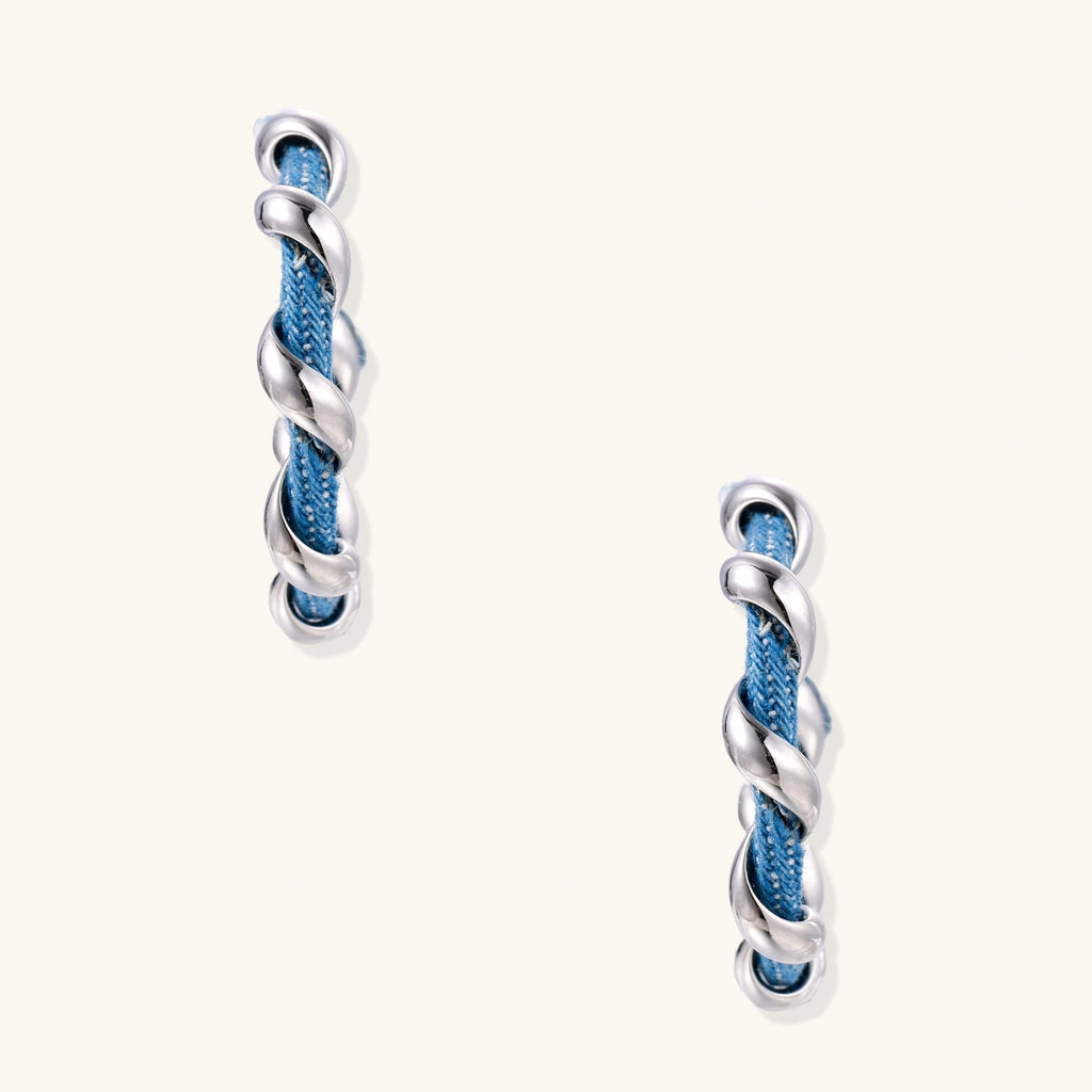 White gold-plated hoops intertwined with blue denim fabric in a twisted rope design on cream background.