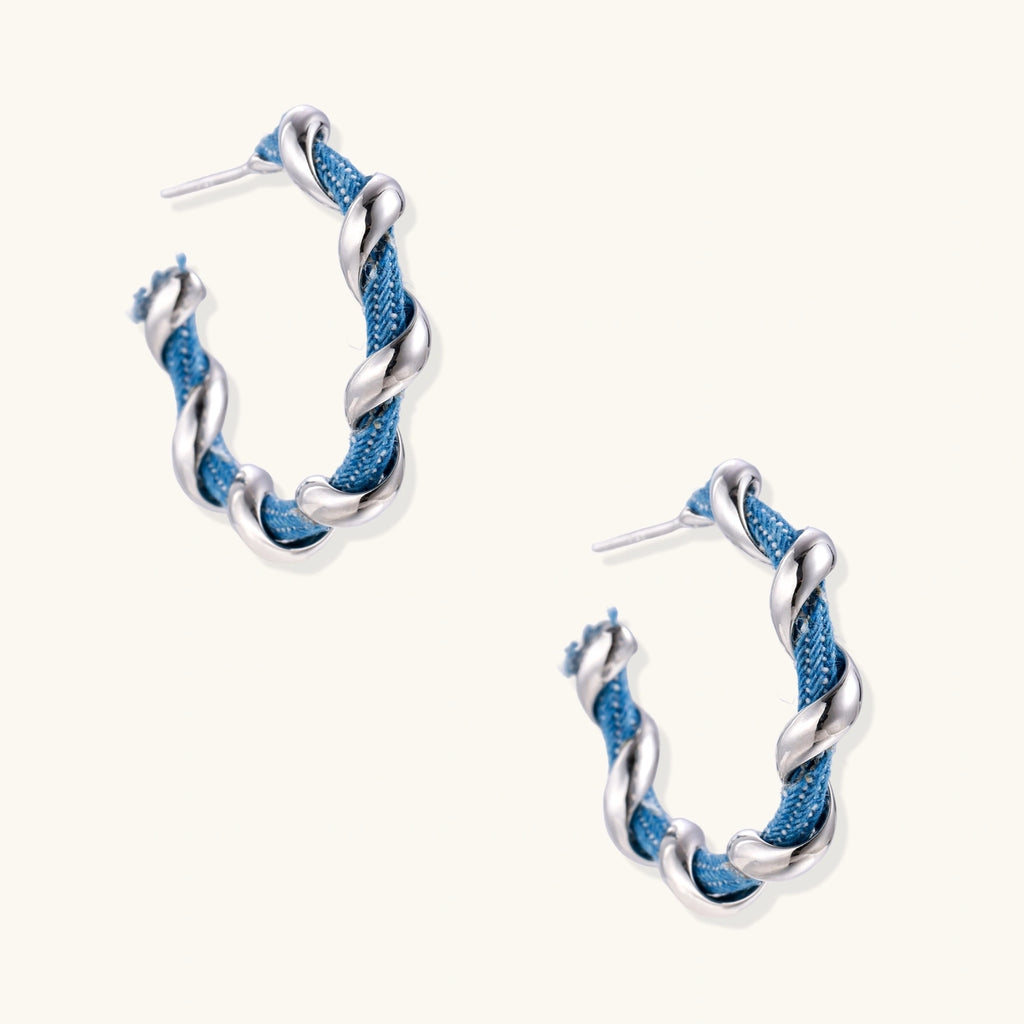 White gold-plated hoops intertwined with blue denim fabric in a twisted rope design on cream background.