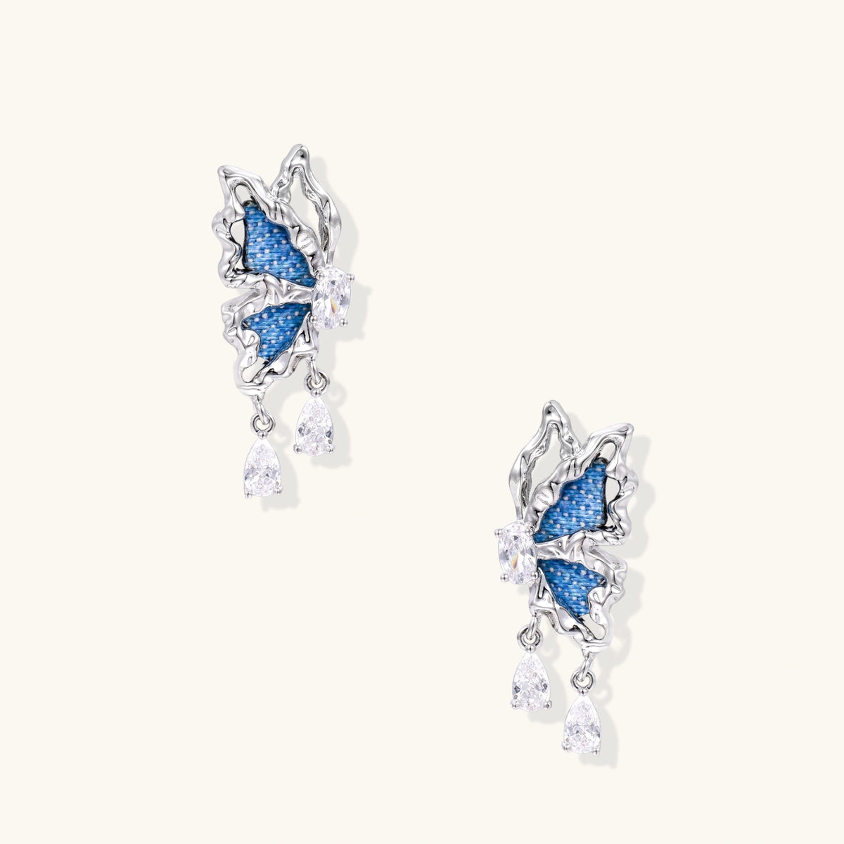 Silver butterfly earrings with blue denim fabric wings and dangling teardrop crystals on cream background